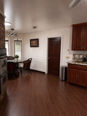 A kitchen with wooden cabinets, a dining table with chairs, a built-in oven, and a trash can. - Spacious Getaway : 4BR-10Guest House Near AT&T Stadium, DFW Airport & Stockyards (Fort Worth)