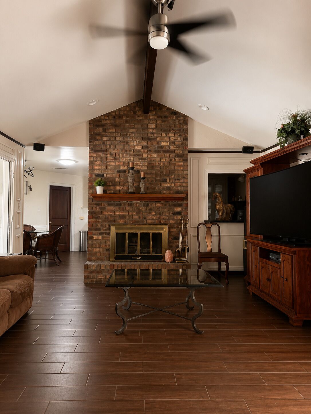A living room with a brick fireplace, a ceiling fan, a flat-screen TV, and a dining area with chairs and a table.