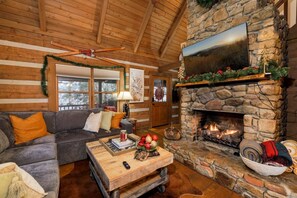 A cozy log cabin interior with a stone fireplace, a couch, and a television mounted above it. - Boone & Banner Retreat🏡.  Private Home. Hot Tub & Fire Pit. Blue Ridge views! (Banner Elk)