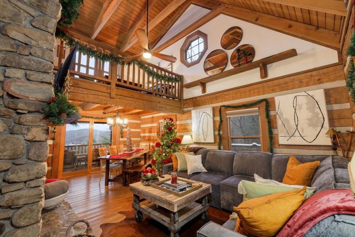 A cozy cabin interior with a stone wall, wooden ceiling beams, a dining area, and a living room with a fireplace.