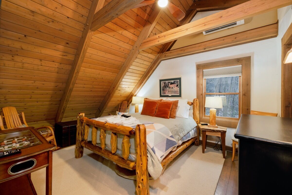 A cozy attic bedroom with a wooden ceiling, a bed with a quilt, a desk with a computer, and a window with a view of trees.