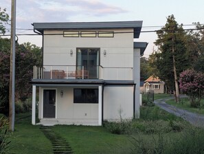 Una casa moderna de dos pisos con balcón, puerta principal y una escalera visible que lleva a la entrada.