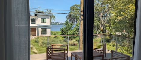 A balcony with wooden furniture, a view of a lake, and a modern building across the water.