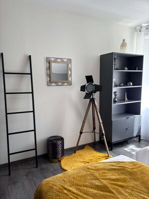 Una habitación con una escalera, un trípode con una cámara, un espejo, una estantería y una alfombra amarilla.