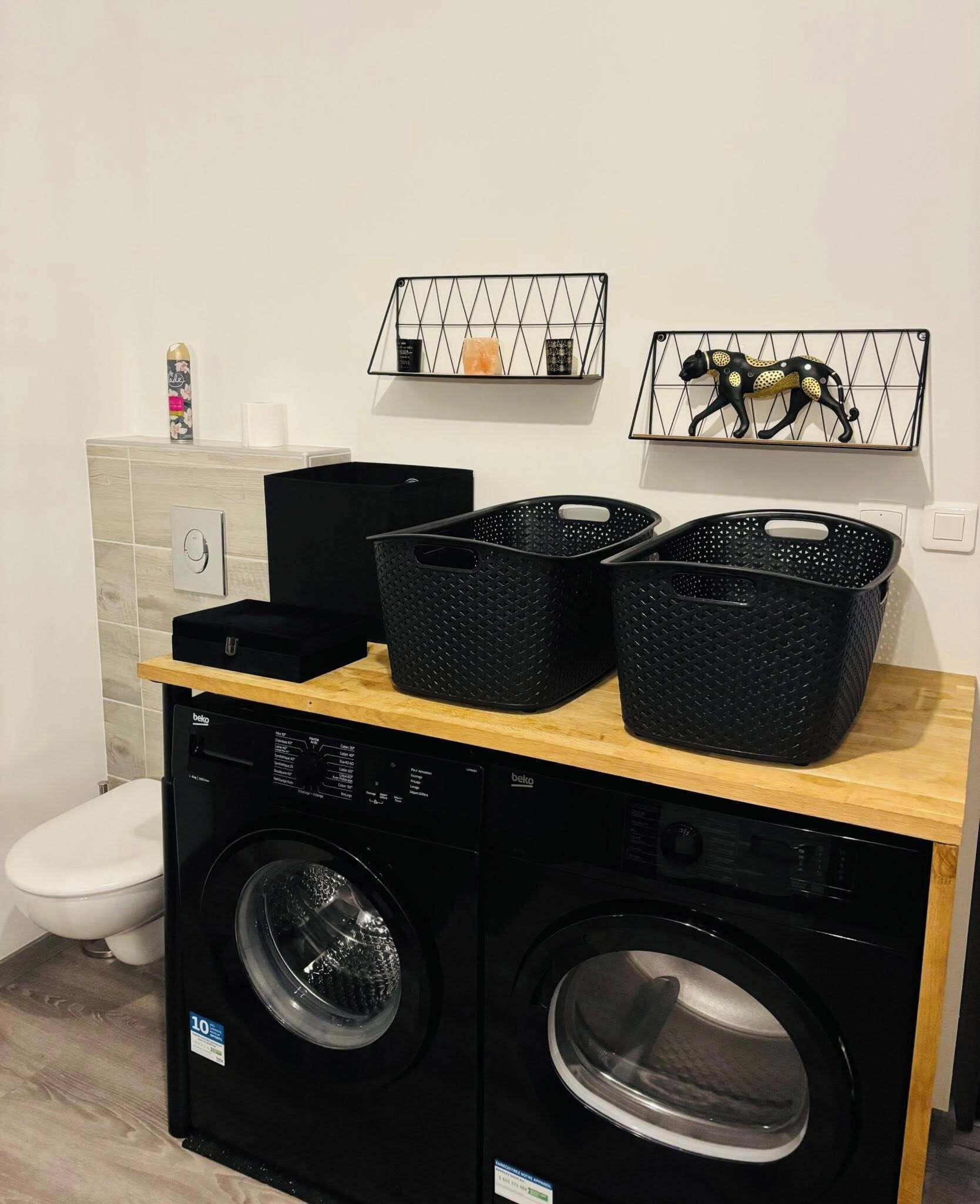 A compact laundry area with a washing machine and dryer, two black baskets, a shelf with decorative items, and a toilet.