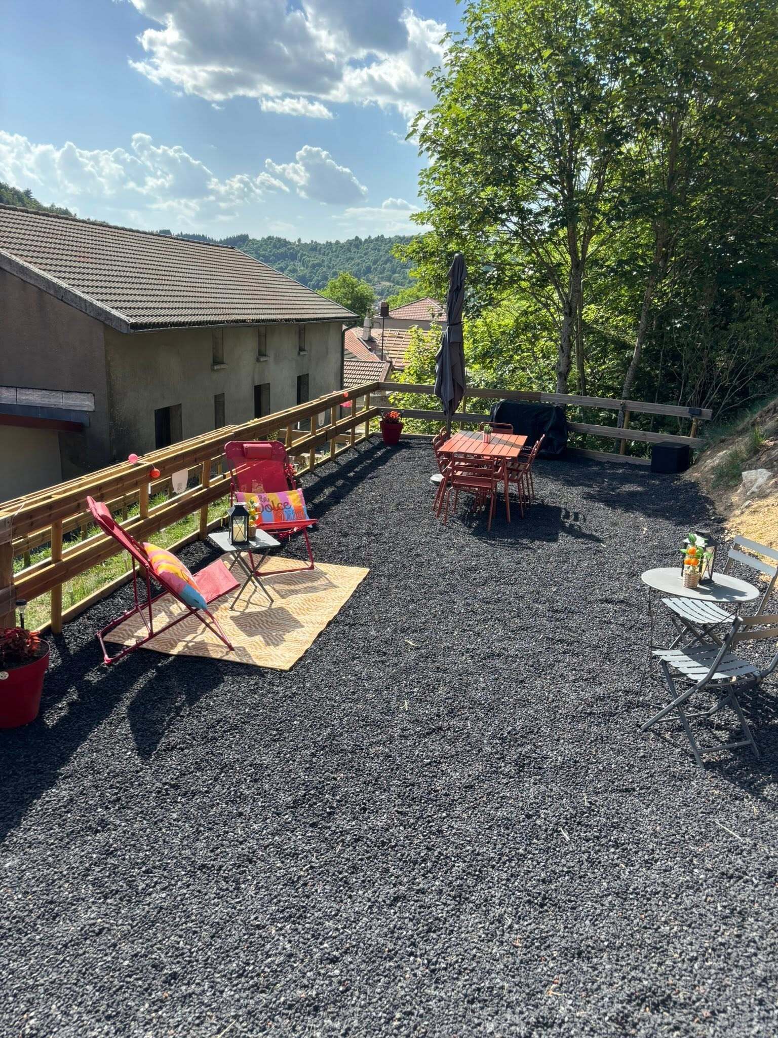 An outdoor seating area with a gravel surface, wooden deck, and a view of a building and trees.
