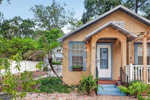 A single-story house with a brick entrance, a small porch with string lights, and a white picket fence. - King Davis Cottage | Fenced In, Downtown (St. Augustine)