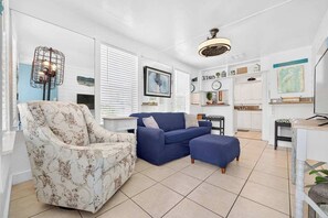 A living room with a patterned armchair, a blue sofa, and a small ottoman. There is a dining area with a table and chairs, and a kitchen area with white cabinets and shelves. - King Davis Cottage | Fenced In, Downtown (St. Augustine)