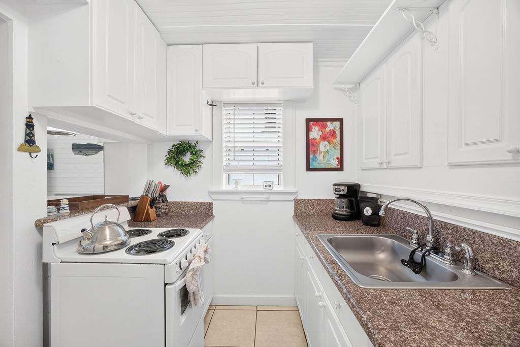A compact kitchen with white cabinetry, a stove, a sink, and a window with blinds.