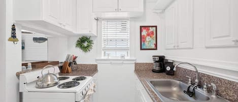 A compact kitchen with white cabinetry, a stove, a sink, and a window with blinds.