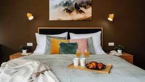 Un dormitorio con cama, almohadas, mesitas de noche, una bandeja con croissants y bebidas, y un cuadro en la pared.