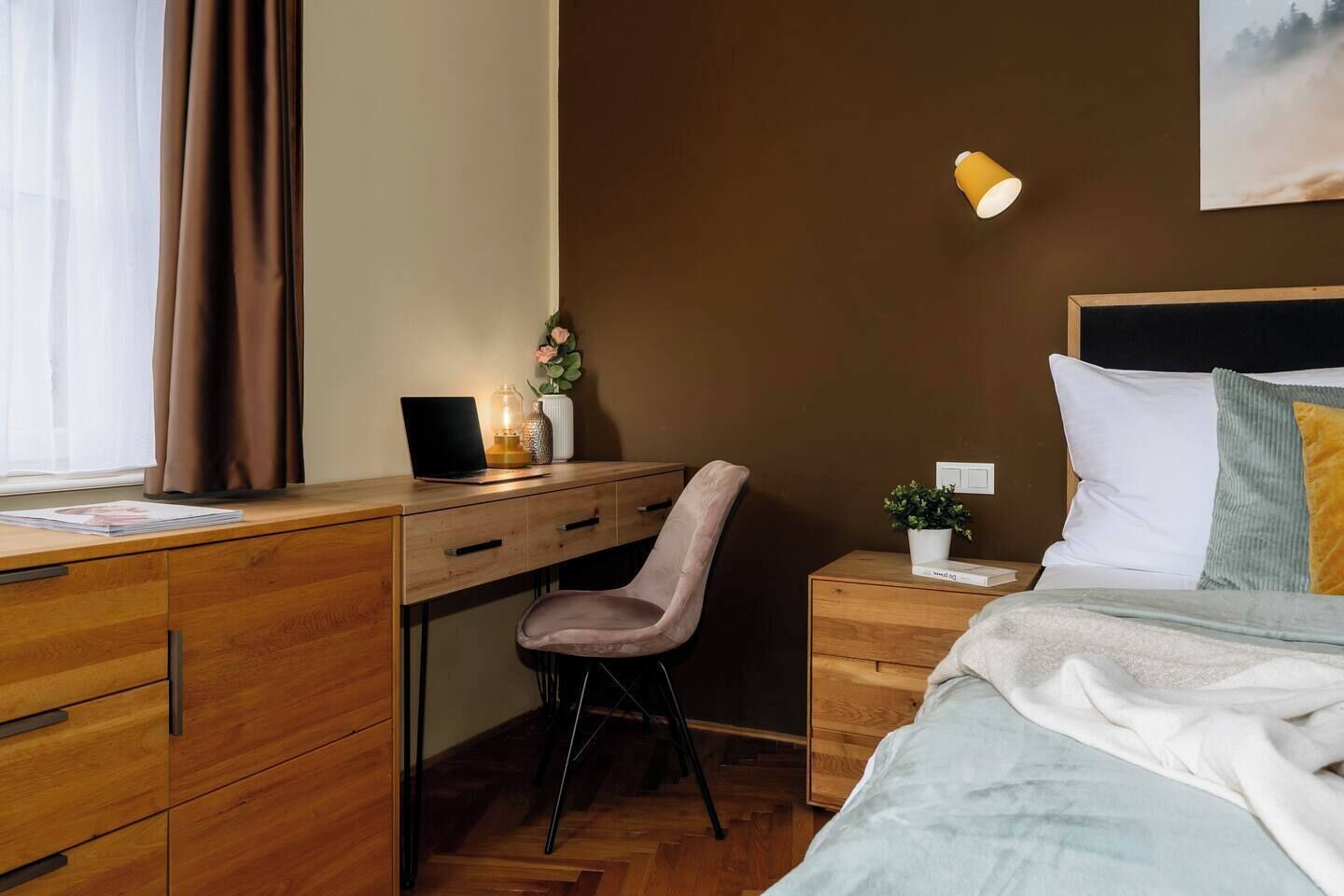 A hotel room with a wooden desk, a chair, a bed with a white headboard, a bedside table with a lamp, and a wall-mounted light.
