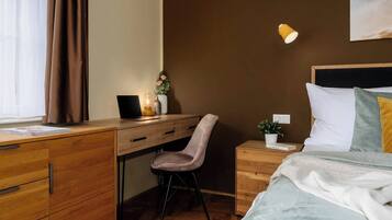 A hotel room with a wooden desk, a chair, a bed with a white headboard, a bedside table with a lamp, and a wall-mounted light.