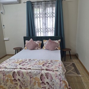Un dormitorio con cama, dos mesitas de noche, una ventana con cortinas y aire acondicionado instalado en la pared.
