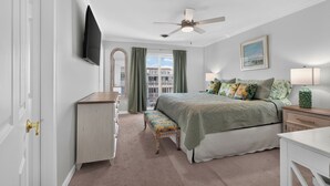 Quarto com cama, escrivaninha, cadeira, televisão, ventoinha no teto e janela com cortinas.