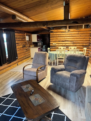 Interior de una cabaña de troncos con comedor, dos sillones, una mesita de centro y una cocina con microondas.