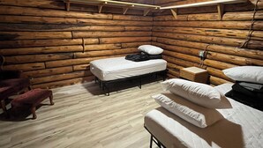 Una habitación tipo cabaña de troncos con dos camas, piso de madera y techo inclinado con una lámpara.