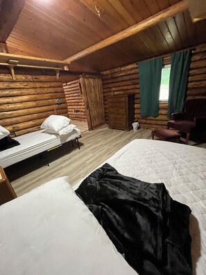 A cabin with two beds, wooden walls, a window with curtains, and a chair. - Charming 2-bedroom log cabin with AC by Lake Metigoshe (Bottineau)
