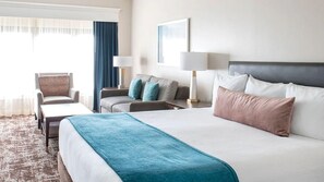 Une chambre d’hôtel avec un grand lit, un canapé, un fauteuil et un bureau. Il y a deux lampes et un tableau au mur.