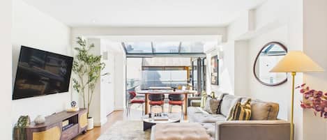 A modern living room with a flat-screen TV, a sofa, a coffee table, and a dining area with chairs and a table.