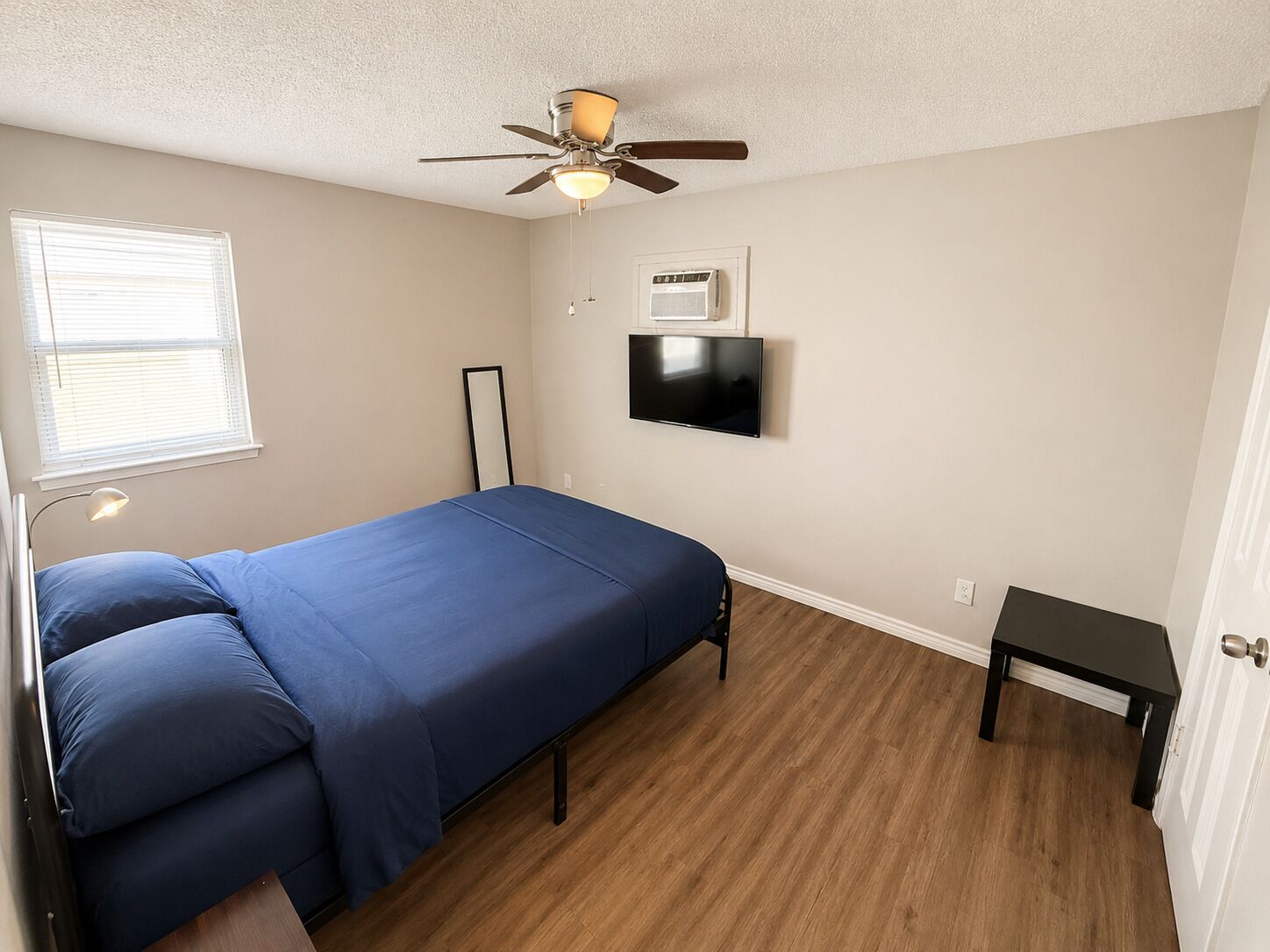 A bedroom with a bed, a ceiling fan, a TV, a window with blinds, and a bedside table.