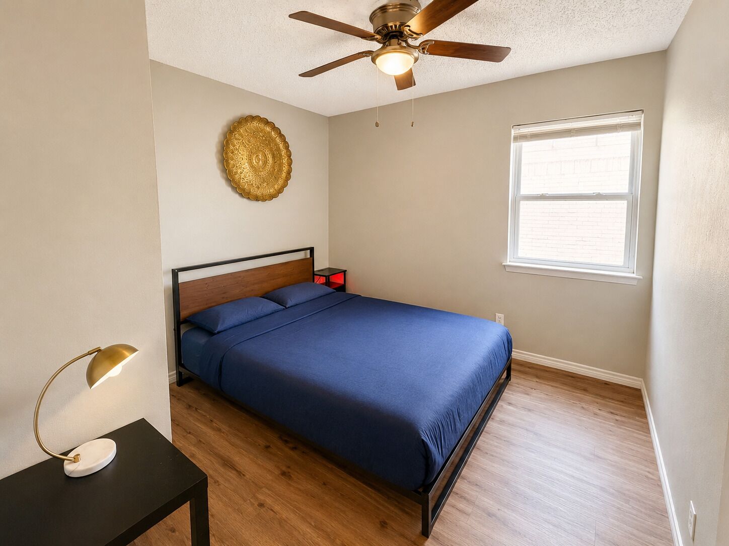 Une chambre à coucher comprenant un lit, un ventilateur de plafond, une assiette décorative fixée au mur, une table de chevet avec une lampe et une fenêtre avec des stores.