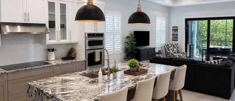 A modern kitchen with a marble island, pendant lighting, and a dining area with a black sofa.