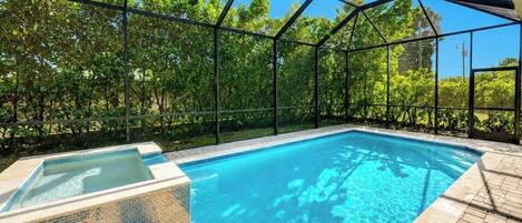 An outdoor swimming pool with a glass enclosure, surrounded by a brick patio and lush greenery.