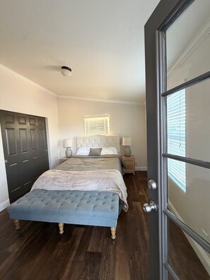 A bedroom with a bed, bedside tables, a window with blinds, a door, and a ceiling light. - Spacious 4 Bedroom home on an 80 acre horse ranch- 15 minutes from Wine Country (Hemet)