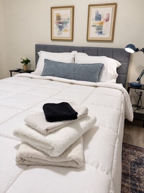Una cama bien hecha con edredón blanco, almohada gris y manta negra. Dos obras de arte enmarcadas en la pared sobre la cama.