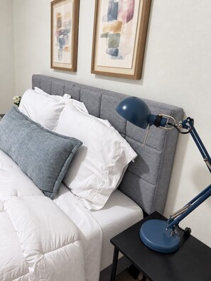 A neatly made bed with white and grey bedding, a blue desk lamp, and two abstract paintings on the wall. - 204 Charming Studio, DT Indy, Medium & Long Stays! (Indianapolis)
