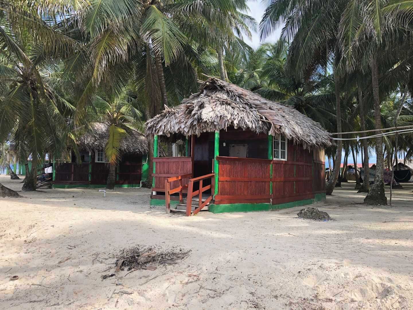 A thatched-roof hut with a wooden porch, surrounded by palm trees on a sandy beach.