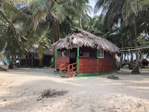 Une hutte à toit de chaume avec une véranda en bois, entourée de palmiers sur une plage de sable.