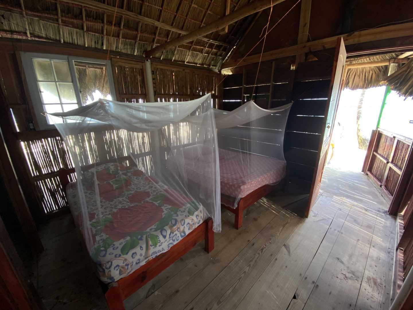 A room with a bed under a mosquito net, wooden walls, and a thatched roof.