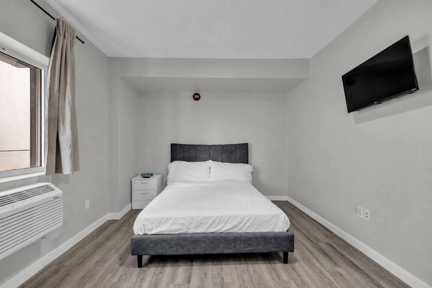 A bedroom with a bed, a nightstand, a wall-mounted TV, and a window with curtains.