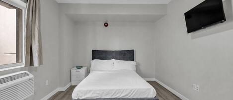A bedroom with a bed, a nightstand, a wall-mounted TV, and a window with curtains.