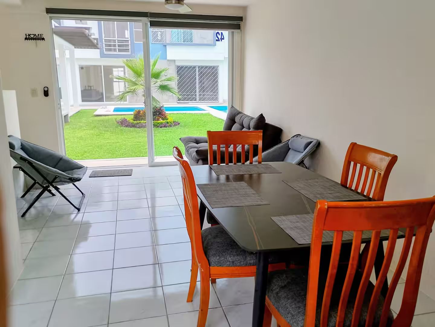 Un coin repas avec une table et des chaises, une porte-fenêtre coulissante donnant sur une piscine, et une pancarte indiquant « MAISON ».