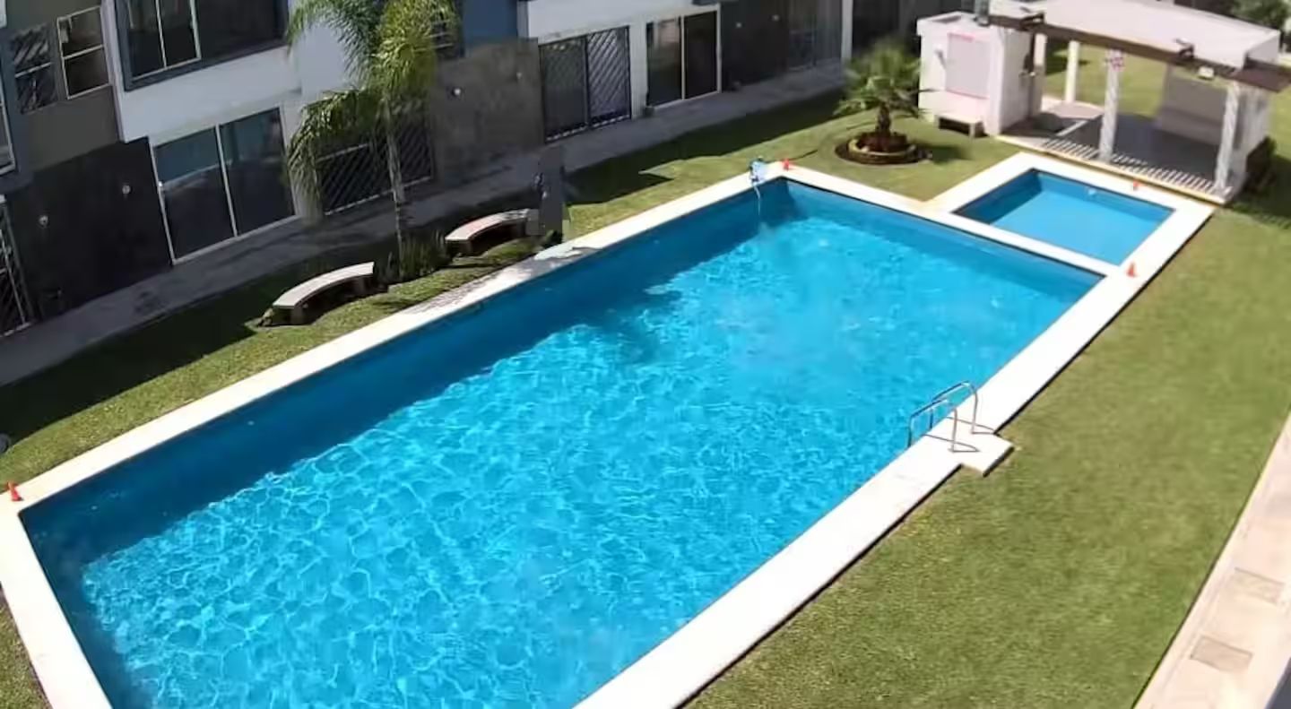 Une piscine rectangulaire bordée de blanc, entourée d’une pelouse bien entretenue et, au fond, d’un bâtiment doté de grandes fenêtres.
