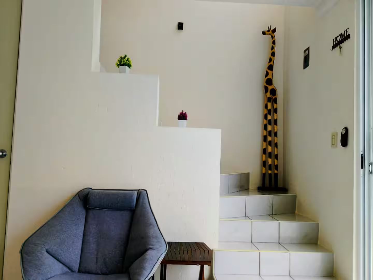 Una stanza con una poltrona blu, una scultura a forma di giraffa e una scala con un cartello che riporta la scritta 'HOME'.