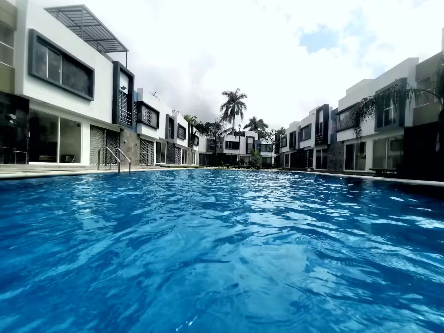 En swimmingpool med moderne bygninger på hver side, en klar himmel og palmer i baggrunden.