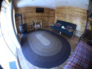 Ein Interieur einer Hütte mit einem runden Teppich, einer Couch, einem Tisch und einem an der Wand befestigten Fernseher.