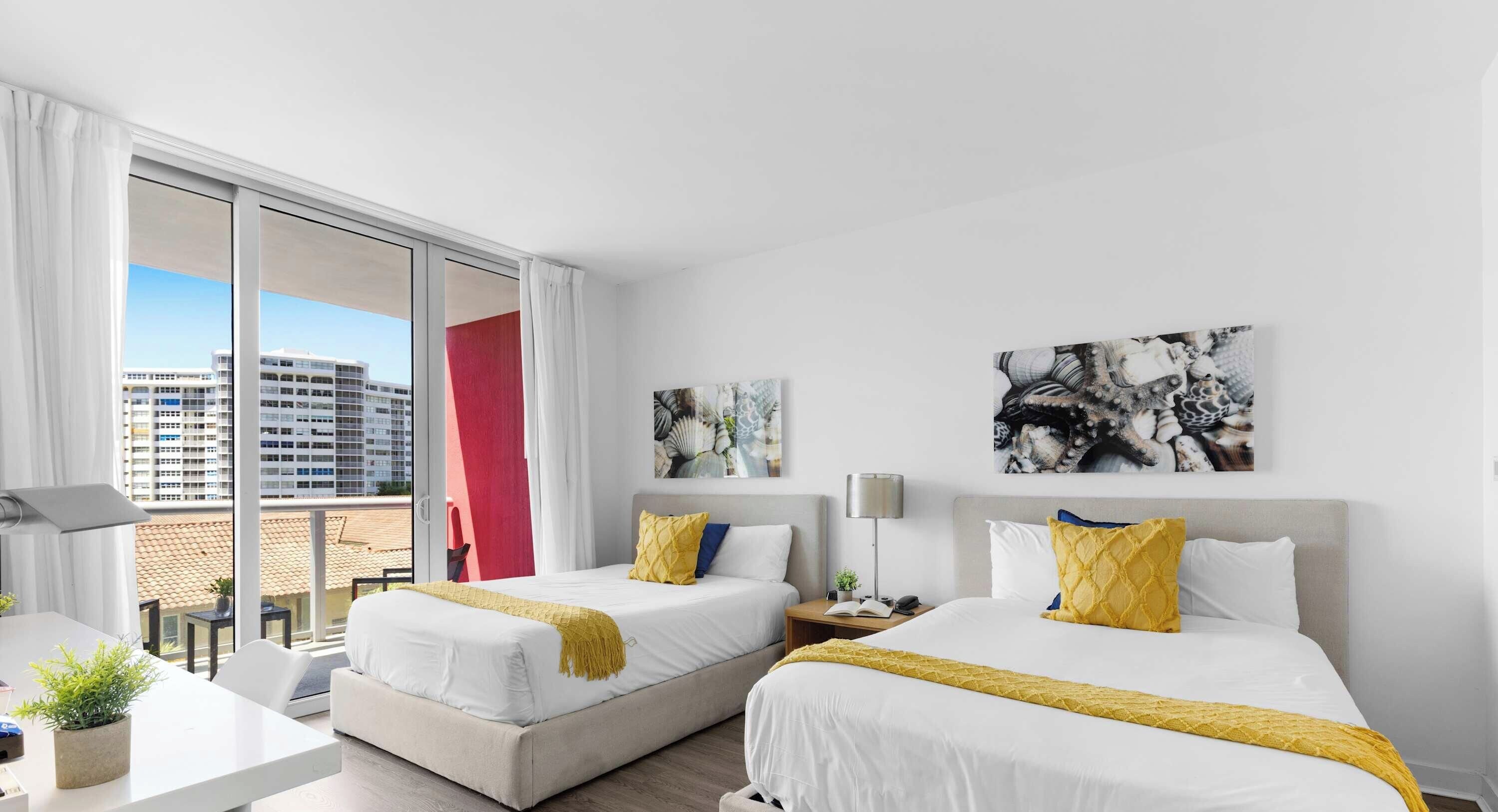 Quarto de hotel com duas camas, uma escrivaninha, uma cadeira e vista para um edifício através de uma grande janela.