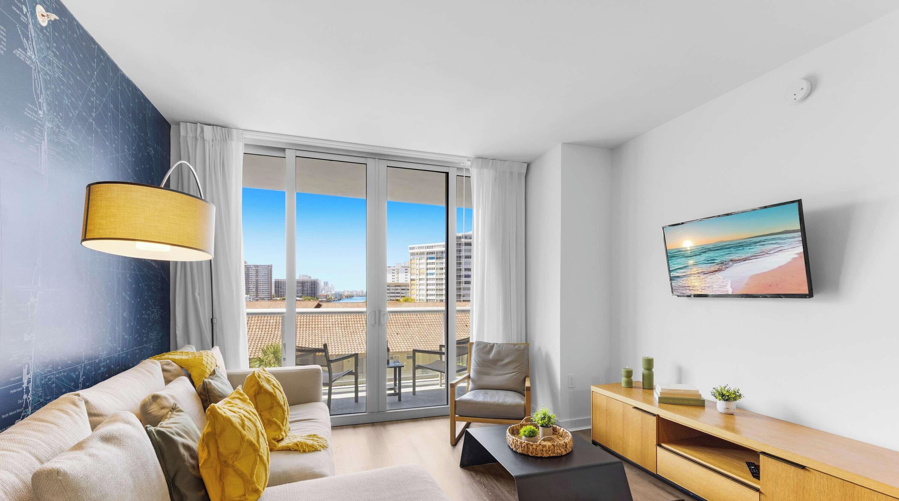 A modern living room with a sofa, a coffee table, a chair, a TV on the wall, and a view of the city through large windows.