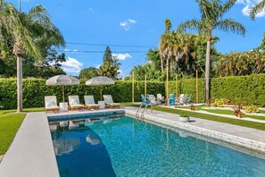 A swimming pool with lounge chairs and umbrellas, surrounded by greenery and palm trees. - Elegant 5BR Retreat- Pickleball + Mini golf + Pool (Bradenton)