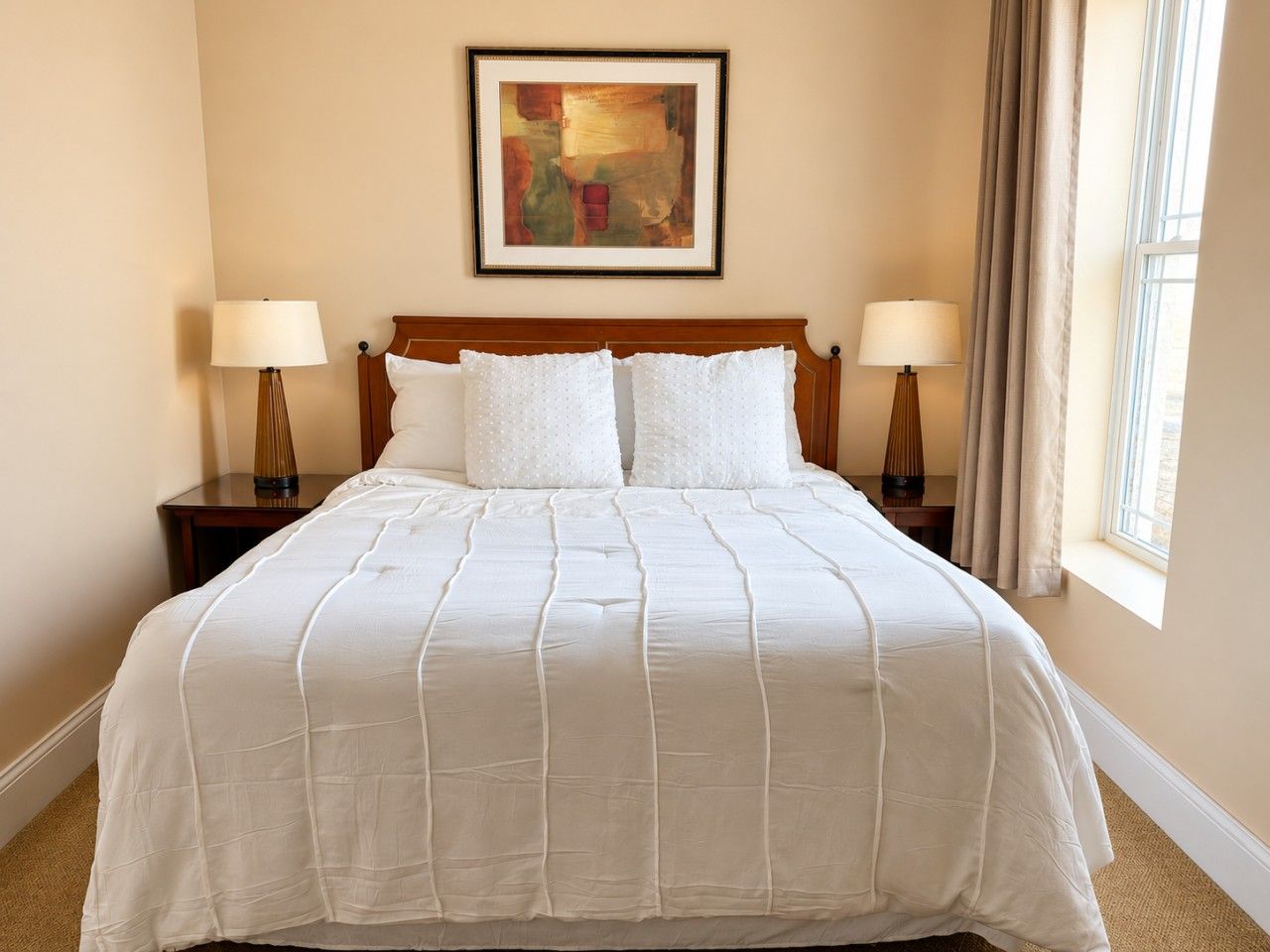 Camera d'albergo con un letto grande, due comodini con lampade, un quadro incorniciato al muro e una finestra con tende.