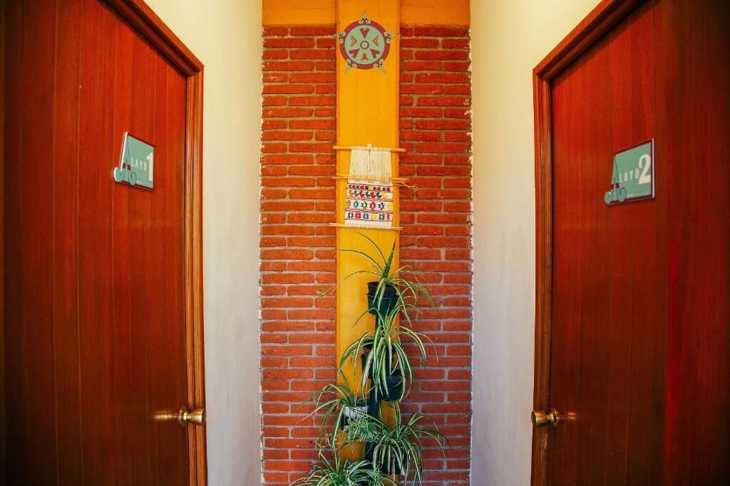 Un pasillo con dos puertas de madera, cada una con un número de habitación. Una columna amarilla vertical con un reloj y una pequeña señal está entre las puertas.