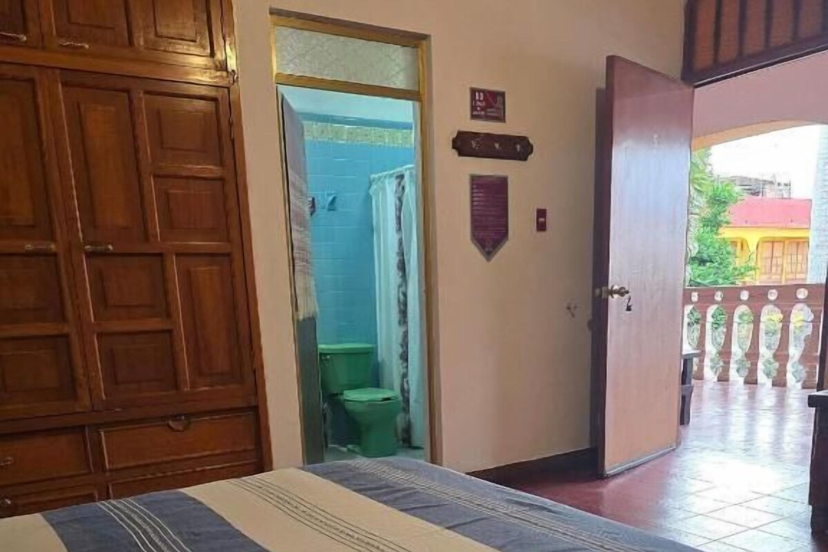 A room with a bed, a wooden door, a bathroom visible through an open door, and a balcony with a railing.