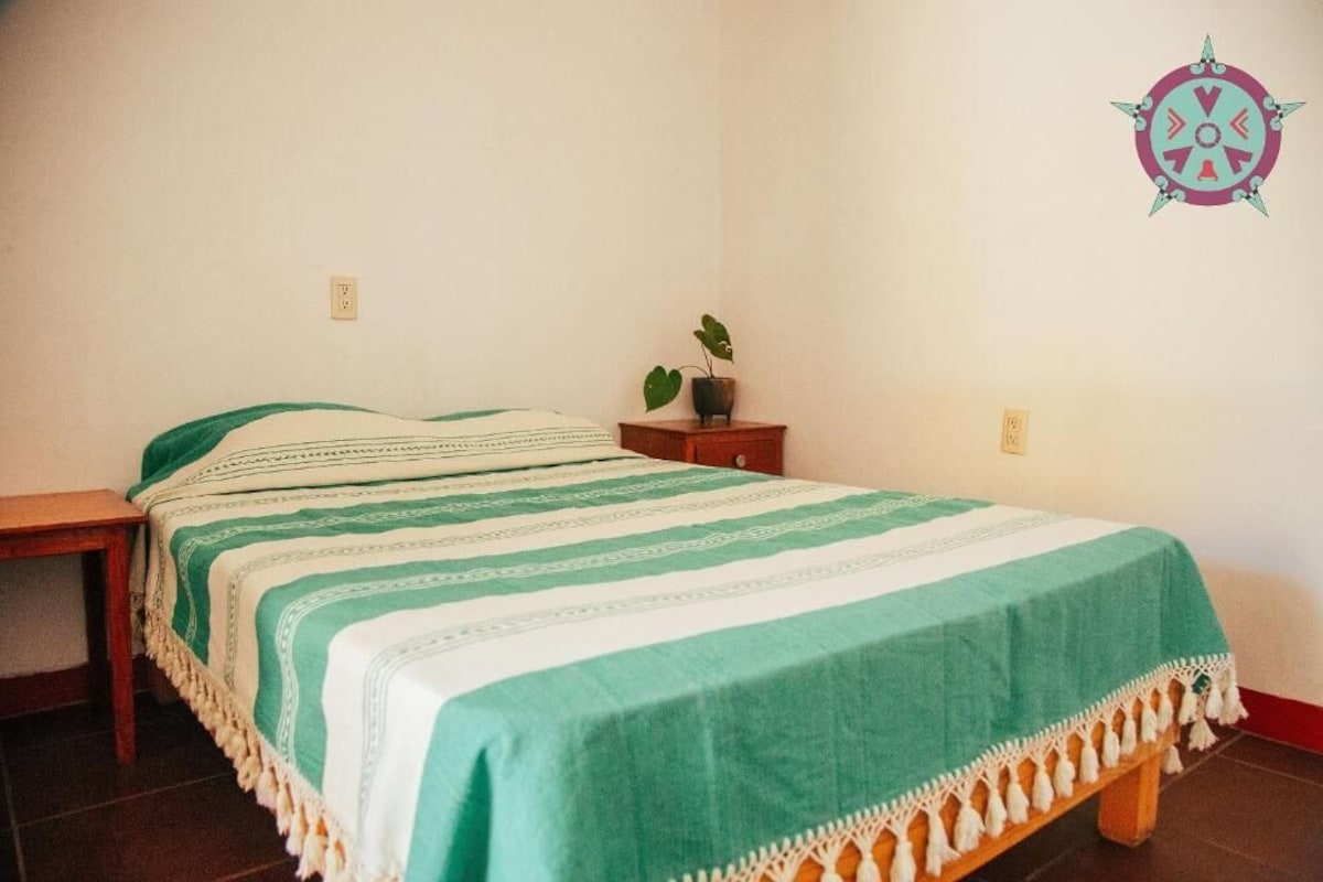 A single bed with a green and white striped bedspread, a bedside table with a plant, and a wall decoration.