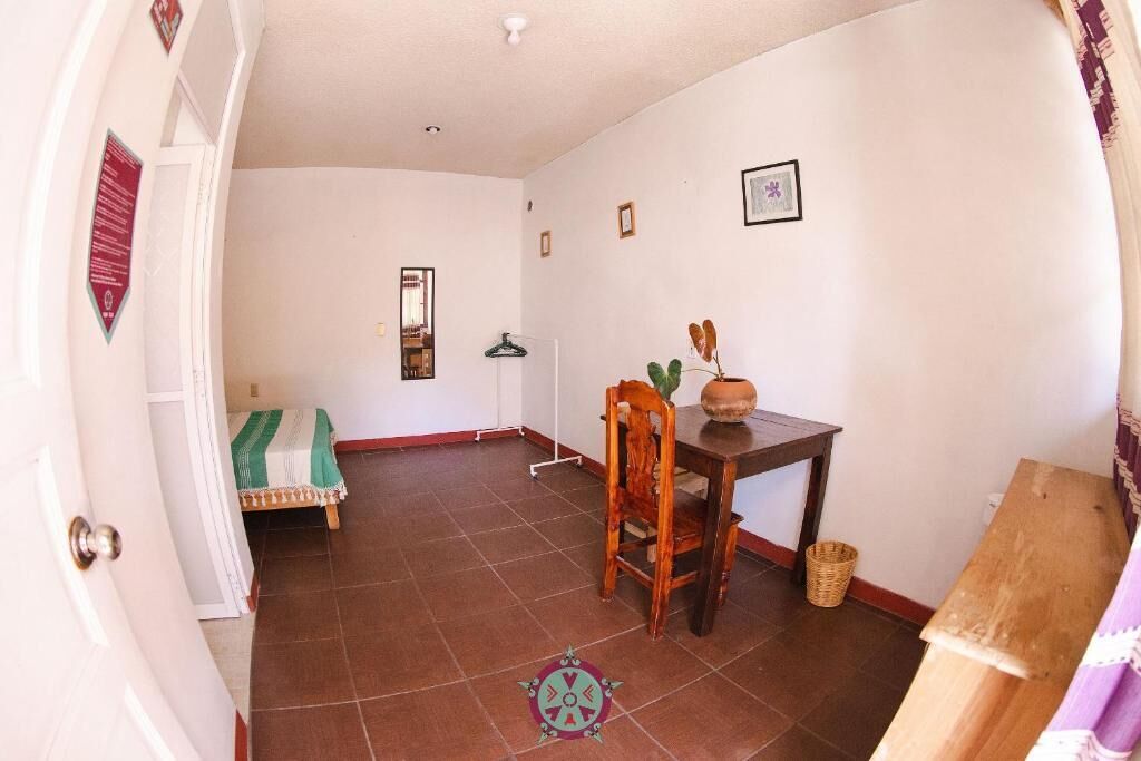 Una habitación con una mesa de comedor, una silla, una mesita con una planta, un perchero y una cama con una colcha verde y blanca a rayas.