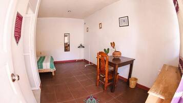Una habitación con una mesa de comedor, una silla, una mesita con una planta, un perchero y una cama con una colcha verde y blanca a rayas.